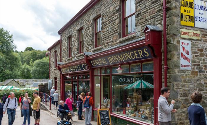 this shows an old shop building made of stone. There are two large shop windows with dark red window frames. you can see Welsh food displayed in one w